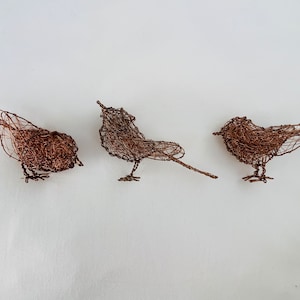 Op de afbeelding: Drie kleine, ingewikkeld geweven koperdraadvogels. De vogels zijn in een rij gerangschikt, met de middelste vogel naar voren gericht en de andere twee naar de zijkant.