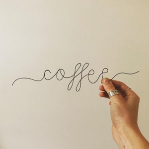 Può includere: Scultura in filo nero che scrive la parola "coffee" su sfondo bianco. La scultura è tenuta da una mano che indossa anelli d'argento. La parola è scritta in una linea continua.
