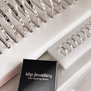 Könnte beinhalten: Eine Sammlung silberner Armreifen mit graviertem Schriftzug, auf einer weißen Oberfläche ausgestellt. Eine schwarze Box mit dem Text "ichu jewellery 925 Sterling Silver" befindet sich im Vordergrund.
