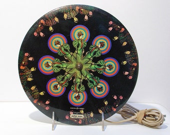 May include: A vintage Peter Max designed decorative plate with a black background. The plate features a central starburst design with figures in green dresses and colorful concentric circles. The plate has a power cord and is displayed on a clear stand.