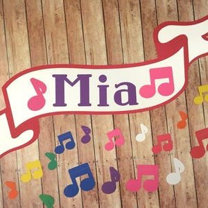 May include: A white banner with red trim and the name "Mia" in purple letters. The banner has two pink musical notes on either side of the name. The banner is hanging on a wood background with several colorful musical notes scattered around it.