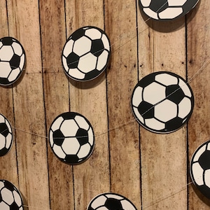 May include: A string of black and white soccer ball cutouts on a wood background.