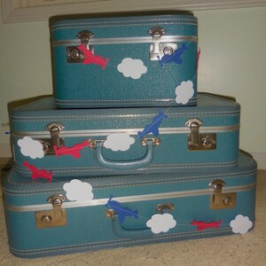 Pode incluir: Tr&ecirc;s malas vintage azul-petr&oacute;leo empilhadas umas sobre as outras. As malas t&ecirc;m detalhes prateados e s&atilde;o decoradas com uma guirlanda de papel de avi&otilde;es vermelhos e azuis e nuvens brancas.