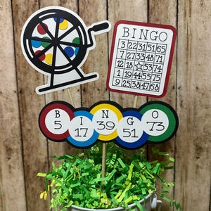 May include: A festive bingo-themed centerpiece featuring a bingo wheel, a bingo card, and bingo number circles. The wheel is black and white with colorful accents. The bingo card has a red border and numbers. The number circles spell BINGO.