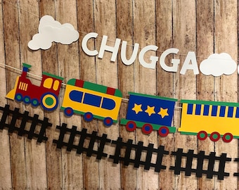 May include: A colorful train banner with the word "CHUGGA" in white letters. The train cars are red, yellow, green, and blue, with black train tracks below and white cloud accents. The banner is strung on a white string.