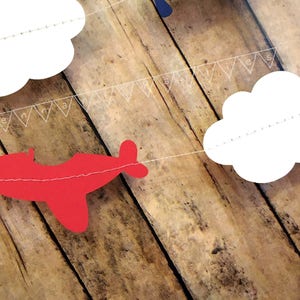 Pode incluir: Um avi&atilde;o de papel vermelho, nuvens de papel branco e uma faixa de papel branco com o texto "Feliz anivers&aacute;rio" s&atilde;o enfiados em um fundo de madeira.