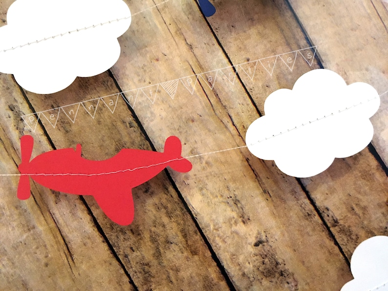Pode incluir: Um avi&atilde;o de papel vermelho, nuvens de papel branco e uma faixa de papel branco com o texto "Feliz anivers&aacute;rio" s&atilde;o enfiados em um fundo de madeira.