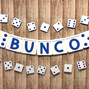 May include: A white banner with blue letters spelling "BUNCO" and white dice with blue dots hanging from it. The banner is hanging on a wood background.