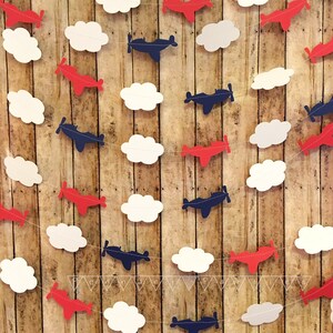 Pode incluir: Uma guirlanda com avi&otilde;es de papel vermelho e azul e nuvens de papel branco. A guirlanda &eacute; enfiada em um barbante branco e est&aacute; pendurada em um fundo de madeira.