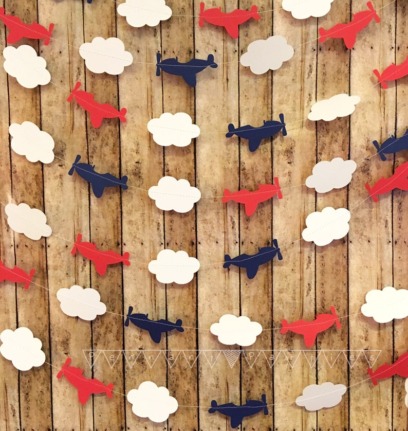 Pode incluir: Uma guirlanda com avi&otilde;es de papel vermelho e azul e nuvens de papel branco. A guirlanda &eacute; enfiada em um barbante branco e est&aacute; pendurada em um fundo de madeira.