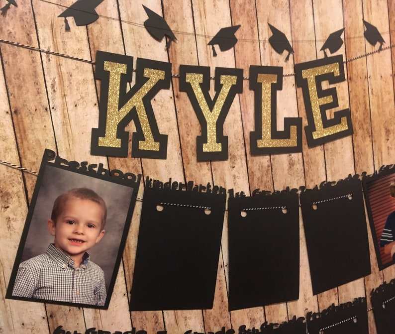 May include: A graduation-themed banner with the name "KYLE" in gold glitter letters on a black background. The banner includes a photo of a child and several black chalkboard-style frames for photos. The background is a wooden plank design.