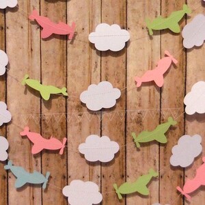 Pode incluir: Uma guirlanda de papel com nuvens brancas e avi&otilde;es em rosa, verde e azul. A guirlanda &eacute; enfiada em um barbante branco e est&aacute; pendurada em um fundo de madeira.