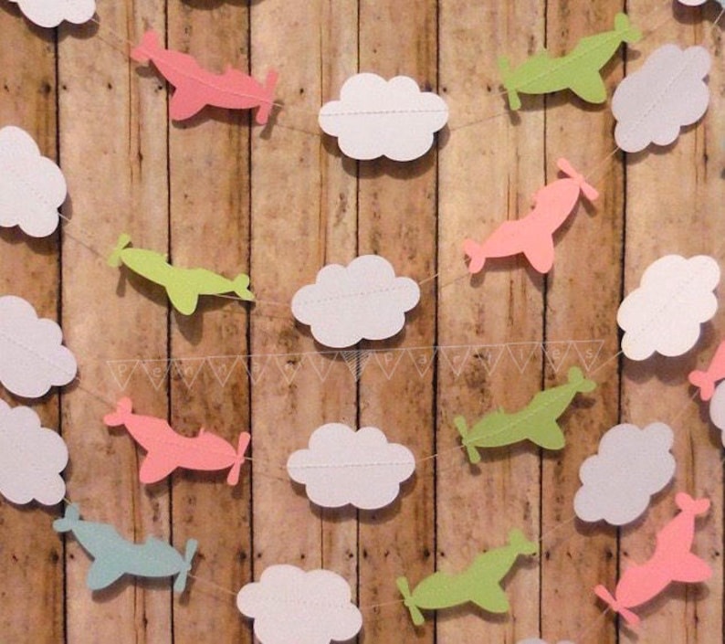 Pode incluir: Uma guirlanda de papel com nuvens brancas e avi&otilde;es em rosa, verde e azul. A guirlanda &eacute; enfiada em um barbante branco e est&aacute; pendurada em um fundo de madeira.