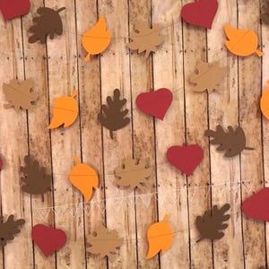 May include: A fall-themed garland with brown, orange, and red paper leaves and hearts. The garland is strung on a white string and is hanging on a wood background.