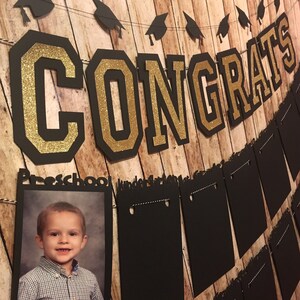 May include: A graduation banner with the word "CONGRATS" in black and gold glitter lettering. A photo of a child in a button-down shirt is displayed below the banner. Additional black pennants and graduation caps are strung across the top.