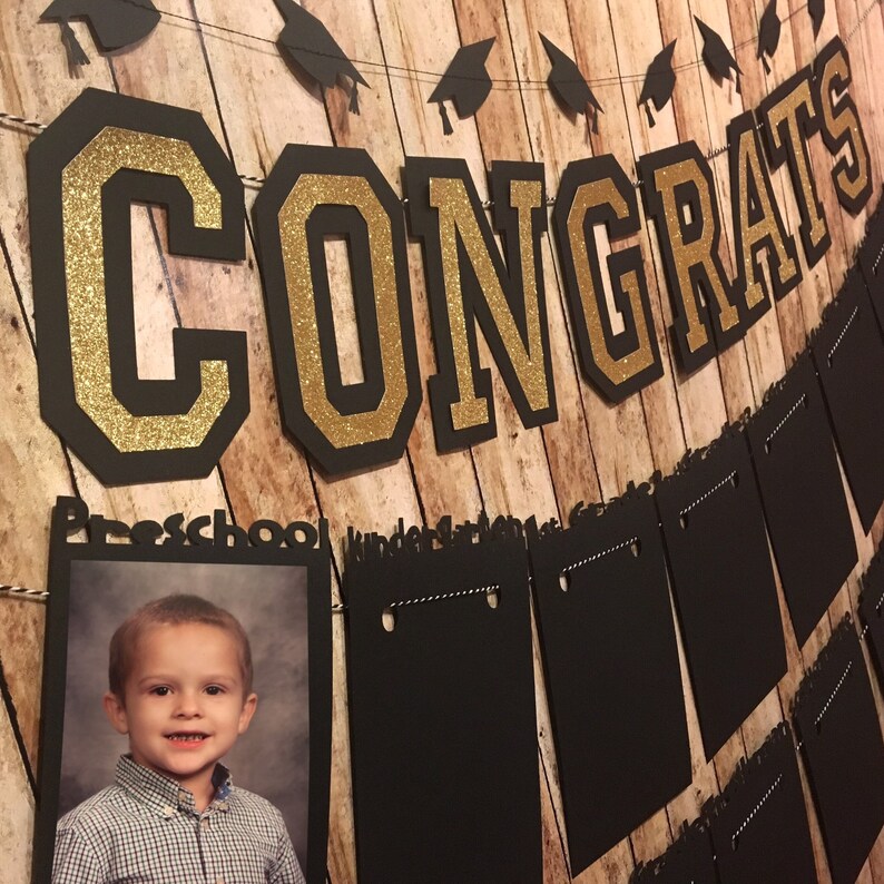 May include: A graduation banner with the word "CONGRATS" in black and gold glitter lettering. A photo of a child in a button-down shirt is displayed below the banner. Additional black pennants and graduation caps are strung across the top.