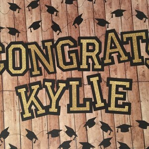 May include: A graduation-themed banner with the words "CONGRATS KYLIE" in gold glitter lettering outlined in black. The background is a wood-grain pattern, and black graduation cap garlands are strung across the top and bottom.