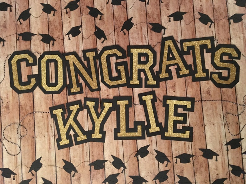 May include: A graduation-themed banner with the words "CONGRATS KYLIE" in gold glitter lettering outlined in black. The background is a wood-grain pattern, and black graduation cap garlands are strung across the top and bottom.