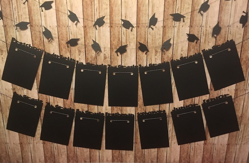 May include: A graduation banner with black mortarboard cutouts strung above black rectangular signs. The signs are labeled with the words "Freshman", "Sophomore", "Junior", and "Senior". The background is a wooden plank wall.