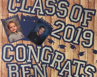 May include: A graduation banner with blue and silver glitter letters that reads "Class of 2019 Congrats Ben". There are also small blue graduation caps scattered around the banner.