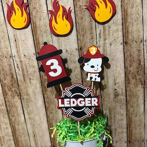 May include: A festive birthday decoration set with a fire department theme. Includes a banner of red and yellow flames, a fire hydrant with the number 3, a Dalmatian firefighter, and a sign that says "LEDGER". The decorations are set in a white bucket with green filler.
