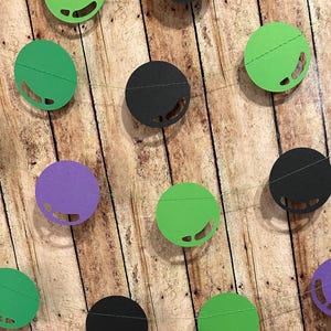 May include: A green, purple, and black paper garland with circles cut out in the shape of a monster's face. The garland is strung on a clear string and is hanging on a wood background.