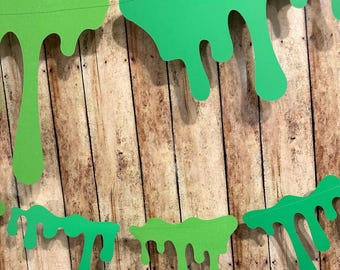 May include: Green paper slime garland with dripping edges. The garland is strung on a white string and is hanging against a wood background.