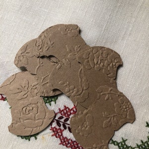 May include: A stack of brown paper gift tags with a floral embossed design. The tags are shaped like a rounded rectangle with a scalloped edge.