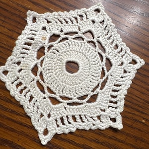 May include: A handmade, off-white crochet doily with a hexagonal shape. The doily features a central circular design surrounded by intricate openwork patterns. The edges are finished with a series of small, decorative loops. The doily is placed on a wooden surface.