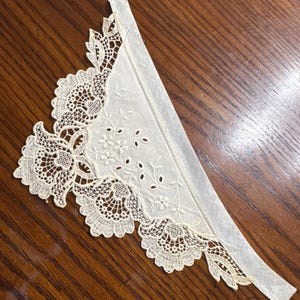 May include: A white lace trim with a scalloped edge and floral designs. The trim is attached to a piece of white fabric.