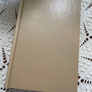 May include: A tan-colored hardcover book with a smooth, slightly textured surface. The book is lying flat on a white crocheted surface.