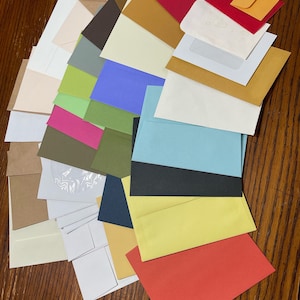 May include: A collection of various colored envelopes and cards, including red, yellow, blue, green, and brown. Some envelopes are small, while others are standard sizes. The items are arranged on a wooden surface.