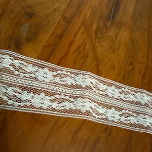 May include: White lace trim with a delicate floral pattern. The trim is made of a fine mesh fabric and has a slightly scalloped edge.