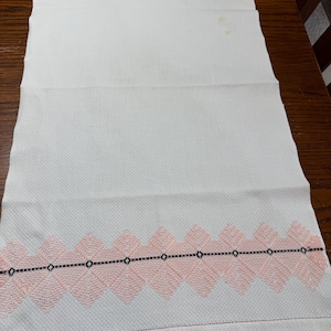May include: White kitchen towel with a pink and black embroidered border. The border features a repeating pattern of diamond shapes.