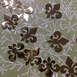 May include: Gold metallic decorative paper cutouts in the shape of fleur-de-lis. The cutouts are scattered on a green and white patterned background.