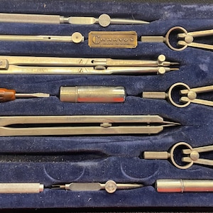 May include: A vintage drafting set in a blue velvet-lined case. The set includes several compasses, dividers, and other drawing tools. The tools are made of metal and have a silver finish. The case is rectangular.