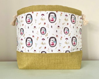 May include: A large, green and white drawstring bag with a pattern of hedgehogs holding apples. The bag is made of a fabric with a hessian texture.