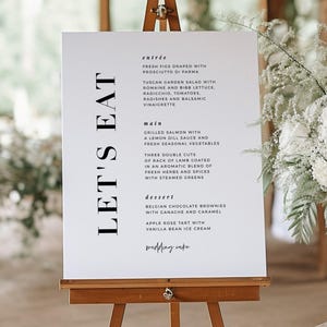 May include: A white sign on a wooden easel with the words "LET'S EAT" in large black letters. The sign lists a menu with entrées, main courses, and desserts, including fresh figs, grilled salmon, and Belgian chocolate brownies.