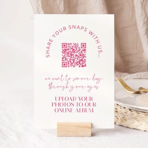 Könnte beinhalten: Eine weiße Karte mit rosa Text und einem QR-Code, gehalten von einem kleinen Holzblock. Der Text lautet "TEILEN SIE IHRE FOTOS MIT UNS..." und "LADEN SIE IHRE FOTOS IN UNSER ONLINE-ALBUM HOCH."