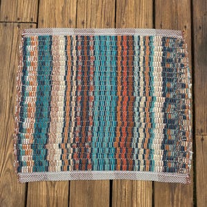 May include: A woven rug with a striped pattern in shades of blue, brown, and white. The rug has a fringed edge.