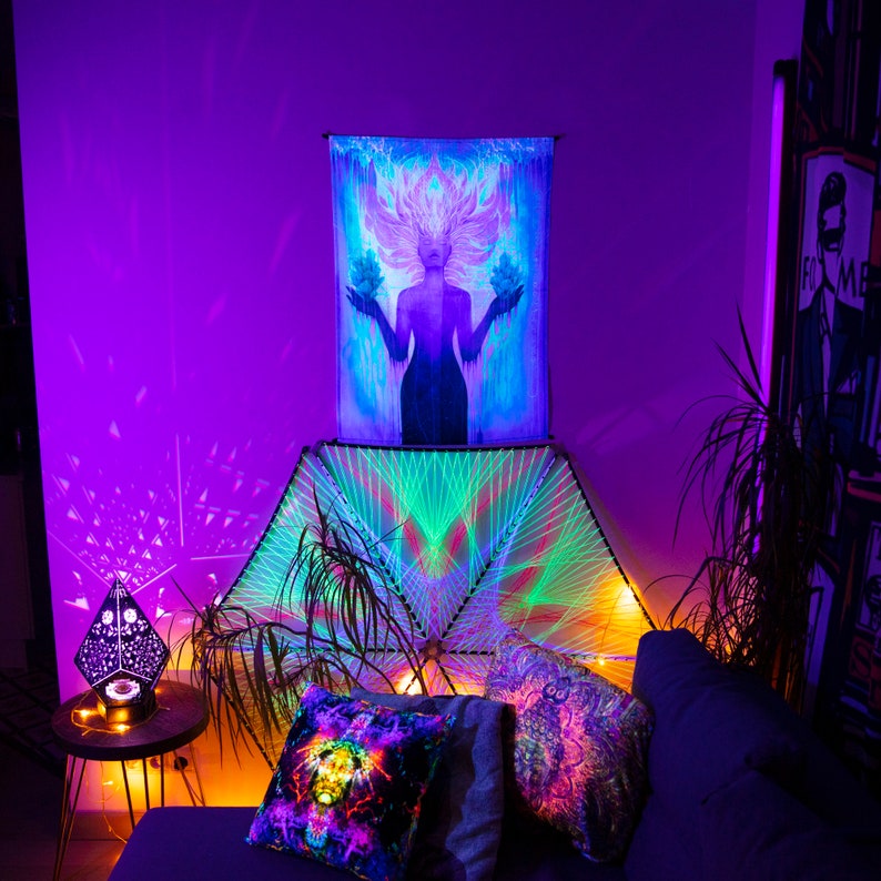 May include: A room decorated with a psychedelic tapestry featuring a woman with flowing hair and a geometric mandala tapestry. The room is lit with colorful lights and features a couch with pillows and a side table with a lamp.