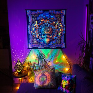 May include: A black tapestry featuring a colourful psychedelic skull design hangs on a purple wall. The tapestry is surrounded by fairy lights and a plant. Two pillows with similar psychedelic designs are on a sofa in front of the tapestry.