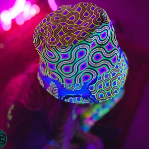 Puede incluir: Un gorro de pescador de color verde neón, azul y morado con un patrón psicodélico de líneas y puntos en espiral. El gorro está hecho de una tela ligera y tiene una visera ancha.