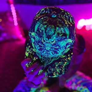 May include: Black baseball cap with a vibrant, intricate neon green and blue design. The cap features a complex pattern of geometric shapes and organic forms, creating a psychedelic effect. The cap is illuminated with a black light.