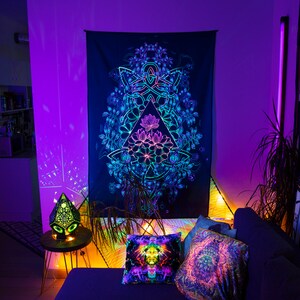 May include: A black tapestry with a colorful mandala design featuring a geometric pattern and floral elements. The tapestry is hanging on a wall in a room with purple and blue lighting.