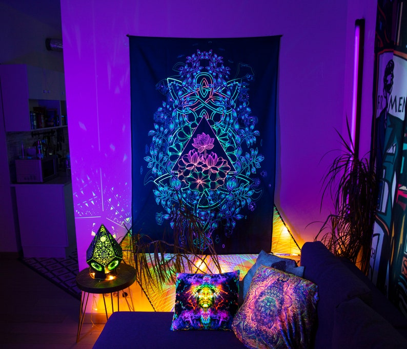 May include: A black tapestry with a colorful mandala design featuring a geometric pattern and floral elements. The tapestry is hanging on a wall in a room with purple and blue lighting.