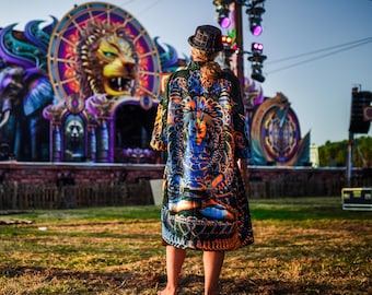 Pode incluir: Uma pessoa usando um quimono estampado com um design detalhado com uma figura de divindade azul. O fundo mostra um palco com uma grande escultura de cabeça de leão e iluminação vibrante, sugerindo um festival ou concerto.