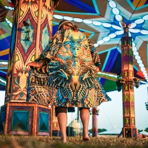 May include: A person wearing a patterned robe stands in front of a decorative structure. The robe features a vibrant mix of blue, orange, and yellow hues. The background includes a large, multi-coloured canopy and ornate pillars.