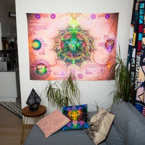 May include: A living room interior with a gray couch, two throw pillows, and a black lamp on a side table. A large tapestry with a colorful mandala design hangs on the wall. A potted plant sits on the floor next to the couch.