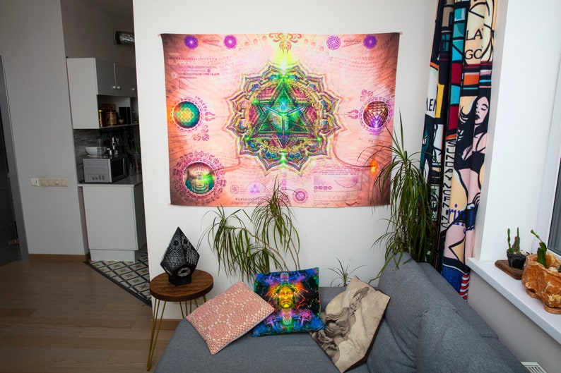 May include: A living room interior with a gray couch, two throw pillows, and a black lamp on a side table. A large tapestry with a colorful mandala design hangs on the wall. A potted plant sits on the floor next to the couch.